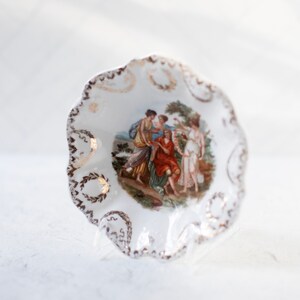 May include: A white porcelain dish with a scalloped edge and gold trim. The dish features a painted scene of four women in a garden setting.
