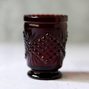 May include: A ruby-red vintage glass tumbler with a diamond and starburst pattern. The rim has a scalloped edge, and the base is a small, circular foot. The glass is likely pressed and has a rich, dark color.