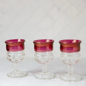 Puede incluir: Tres vasos de chupito de vidrio transparente con un diseño texturizado y un borde de color burdeos.