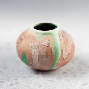 May include: A small, round ceramic vase with a wide opening. The vase features a marbled design in shades of brown, green, and white. The interior of the vase is dark. The vase is likely handmade and suitable for small floral arrangements or decorative purposes.