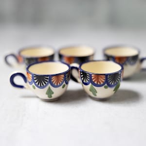 May include: Five small white ceramic cups with blue, green, and orange painted designs. The cups have a rounded shape and a small handle.