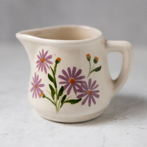 Puede incluir: Una pequeña jarra de cerámica blanquecina con asa y boquilla. La jarra está decorada con flores moradas en forma de margarita con centros naranjas y tallos y hojas verdes. La jarra mide aproximadamente 10 cm de altura.