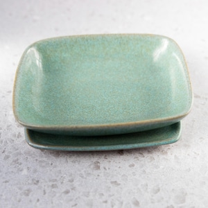 May include: Two turquoise ceramic rectangular plates with rounded corners are stacked on a light gray surface. The plates have a speckled texture and a slightly raised edge with a darker rim.
