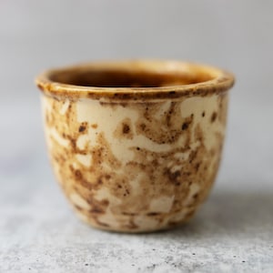 May include: A small, handmade ceramic cup with a cream-colored base and brown, swirling patterns. The rim and interior have a darker brown glaze. The cup has a rounded shape and is likely used for drinking.