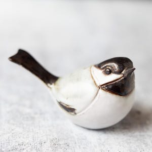 May include: A small ceramic bird figurine featuring a glossy white body with dark brown accents on the head, beak, wings, and tail. The bird is depicted in a resting position with its tail angled upward, showcasing a smooth, glazed finish.