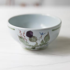 May include: A light blue ceramic bowl featuring a hand-painted botanical design on the exterior. The illustration depicts a dark purple thistle flower, green leaves, and small red berries. A thin green line encircles the upper rim of the bowl.