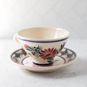 May include: A cream-coloured teacup and saucer set with a floral design. The cup and saucer feature a hand-painted pattern of red, green, and blue flowers and leaves. A blue stripe accents the rim of the cup. Ideal for serving tea.