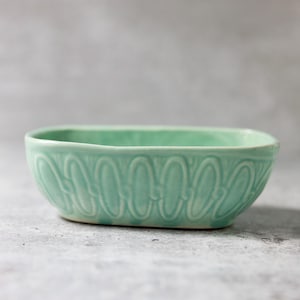 May include: A light green ceramic oval dish with a decorative embossed pattern. The dish is perfect for serving snacks or holding small items.