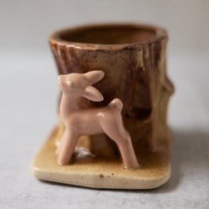 Puede incluir: Un macetero de cerámica con forma de tronco de árbol con una figurita de ciervo rosa unida al lateral. El macetero tiene un esmalte marrón y beige.