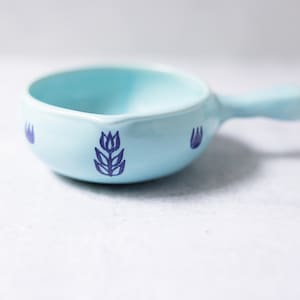 Könnte beinhalten: Eine hellblaue Keramik-Ramekin mit Henkel. Die Ramekin hat ein blaues Blumenmuster auf der Vorder- und Rückseite.