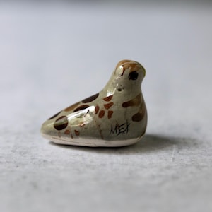 May include: A small, ceramic bird figurine in shades of tan, brown, and cream. The bird has brown spots and a brown head. The base is white, and the word "MEX" is written on the side. A decorative item.