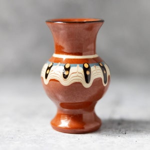 May include: A small, brown ceramic vase with a white and blue design. The vase has a rounded body and a narrow neck.