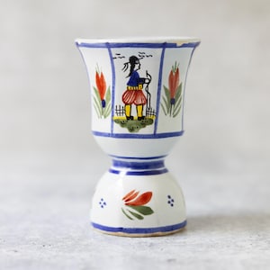 May include: A white ceramic egg cup with a blue rim and base. The cup features a hand-painted design of a person in traditional clothing, red tulips, and a blue border. The egg cup is approximately 10 cm tall.