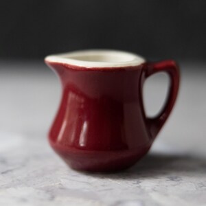 May include: A small, burgundy-colored ceramic creamer with a white rim and a curved handle. The creamer has a glossy finish and a classic, rounded shape. It is designed for serving cream or milk.