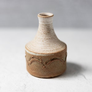 Puede incluir: Un pequeño jarrón de cerámica blanca con una base texturizada marrón. El jarrón tiene un diseño simple y elegante con una abertura ligeramente acampanada y un cuerpo redondeado. Presenta una banda decorativa de líneas marrones en relieve alrededor de la base.