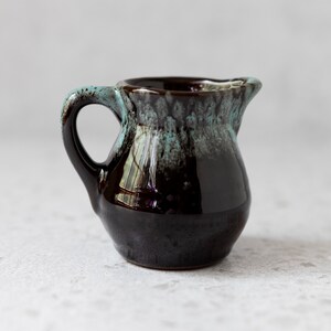 May include: A small, black ceramic creamer with a light blue glaze. The creamer has a rounded body and a handle.