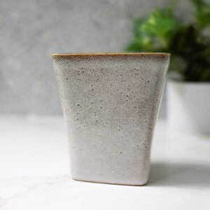 May include: A speckled, off-white ceramic tumbler with a slightly tapered shape. The rim has a brown glaze, and the body has a speckled texture. The mug is designed with a modern aesthetic.