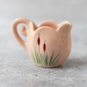 Puede incluir: Un pequeño creamer de cerámica rosa con un diseño floral. El creamer tiene un asa y una forma redondeada. El diseño floral presenta hojas verdes y flores rojas.