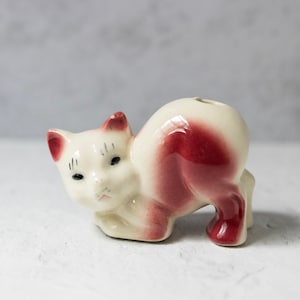 Puede incluir: Una figurita de cerámica de un gato pintada en blanco y rosa. El gato está enroscado en una pose juguetona con la cabeza inclinada hacia un lado. La figurita tiene un pequeño agujero en la parte superior de su espalda, lo que sugiere que puede ser una maceta o un porta lápices.