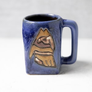 Puede incluir: Una taza de cerámica azul con un asa cuadrada y un diseño único. La taza presenta una imagen marrón y beige de un pájaro o animal. La taza es de color azul cobalto.