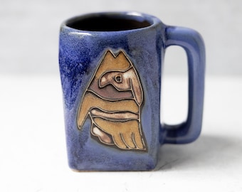 Puede incluir: Una taza de cerámica azul con un asa cuadrada y un diseño único. La taza presenta una imagen marrón y beige de un pájaro o animal. La taza es de color azul cobalto.