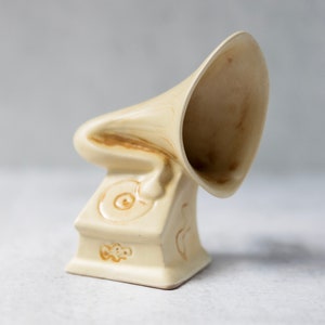 May include: A ceramic figurine of a vintage gramophone with a cream-colored finish and brown accents. The gramophone has a large, round speaker and a small, round record player.