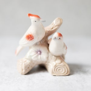 May include: A white porcelain figurine of two birds perched on a branch with a flower. The birds have brown accents on their wings and heads.