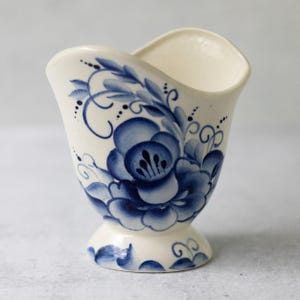 May include: A cream-colored ceramic creamer or small pitcher featuring a hand-painted blue floral pattern. The vessel has a wide, curved rim, a tapered body, and a circular pedestal base, decorated with blue leaves and swirling vine accents.