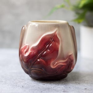 May include: A ceramic vase with a leaf design. The vase has a cream-coloured top and a deep red base, with a gradient of colours in between. The leaves are raised and add texture to the vase.