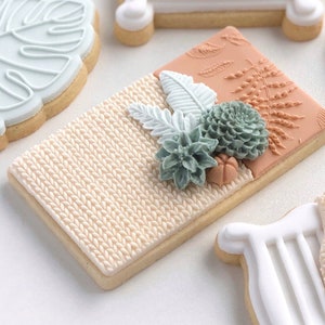 May include: A set of six decorated sugar cookies featuring a cot, a knitted blanket, a monstera leaf, a fern, and succulents. The cookies are decorated with white, light blue, and brown icing.