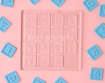Puede incluir: Un molde de plástico transparente con letras y números en relieve, incluyendo el alfabeto y los números 1, 2 y 3. El molde está rodeado de bloques de letras de plástico azul sobre un fondo rosa. El texto "SWEET SPRINKLES! BABY BLOCKS Q-8" está impreso en el molde.
