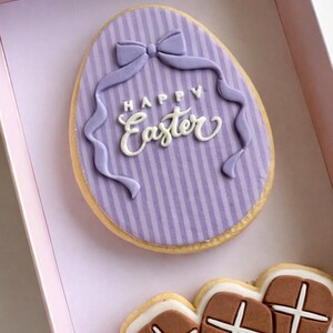 May include: An Easter cookie in a pink box. The egg-shaped cookie has a purple striped background with a bow and ribbon design. The words "Happy Easter" are written in white script. Additional cookies are visible.
