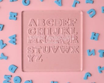 Puede incluir: Un sello de alfabeto de plástico transparente con las letras de la A a la Z en una fuente retro. El sello está sobre un fondo rosa rodeado de letras azules en forma de chispas.