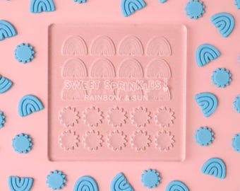 Puede incluir: Un molde de silicona transparente con dos filas de formas de arcoíris y sol. El molde está sobre un fondo rosa con formas de arcoíris y sol azules esparcidas alrededor. El texto "SWEET SPRINKLES! RAINBOW & SUN" está impreso en el molde.