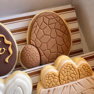 May include: A selection of Easter biscuits, featuring egg shapes with textured patterns and a basket design. The biscuits are decorated with brown, beige, and white icing, presented in a box with striped paper.