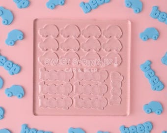 Puede incluir: Un molde de silicona transparente con dos filas de formas de coches y tres filas de la palabra "beep" en una fuente similar a un claxon de coche. El molde está sobre un fondo rosa con formas de coches y "beep" azules esparcidas alrededor.