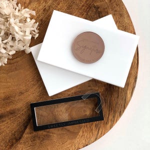 May include: A product shot featuring a brown eyeshadow compact with the word "Signature" in a cursive font, resting on two white rectangular surfaces. A black rectangular box with the text "SWEETLY IMPRESSED" is also visible, all on a wooden surface.