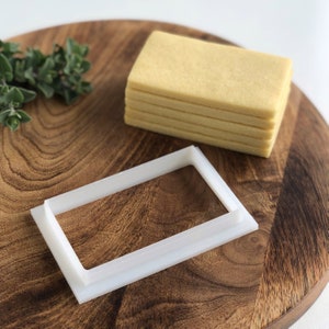 May include: A white plastic rectangular cookie cutter on a wooden surface with a stack of light brown rectangular cookies.