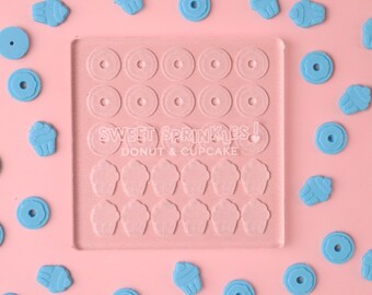 Puede incluir: Un molde de silicona transparente con 16 formas de donut y 16 formas de cupcake. El molde está sobre un fondo rosa con formas de donut y cupcake azules esparcidas alrededor. El molde tiene el texto "Sweet Sprinkles! Donut & Cupcake" impreso en él.