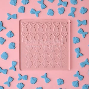 May include: A pink, square mould featuring mermaid tail and seashell designs, with the text "Sweet Sprinkles! Mermaid Tail & Shell". Light blue mermaid tail and seashell-shaped sprinkles are scattered around the mould, set against a pink backdrop.