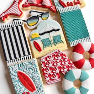 May include: A collection of summer-themed biscuits, including a red bikini top, sunglasses, beach towel, and swim shorts with a lobster pattern. Other biscuits feature a surfboard, crabs, and the text "Adventure Awaits" and "Farewell We'll Miss You".