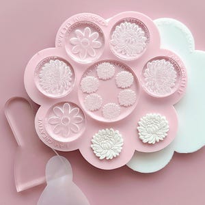 May include: A pink silicone mould with eight different flower designs. The mould is shaped like a flower and has the text "Sweetly Impressed" on the top. The mould is surrounded by a white silicone mat.