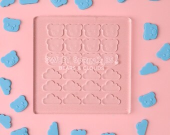 Puede incluir: Un molde rectangular rosa con formas de oso y nube, con el texto "SWEET SPRINKLES! BEARS & CLOUDS". Adornos en forma de oso y nube de color azul claro esparcidos sobre un fondo rosa.
