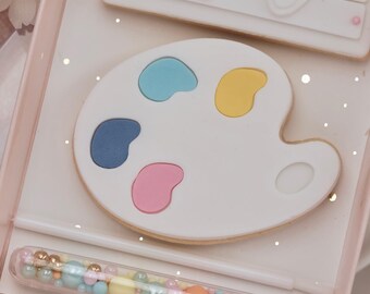 May include: A pink gift box containing a white paint palette cookie, a white cookie with two bunny footprints, and a clear tube filled with colourful sprinkles.
