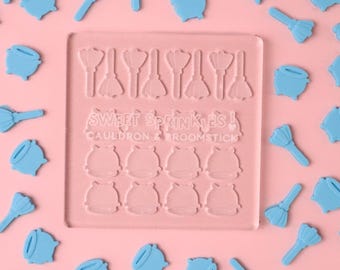 Puede incluir: Un molde de plástico transparente con el texto "Sweet Sprinkles! Cauldron & Broomstick" en él. El molde tiene 12 formas de caldero y 7 formas de escoba. También hay muchas formas de caldero y escoba azules esparcidas sobre un fondo rosa.