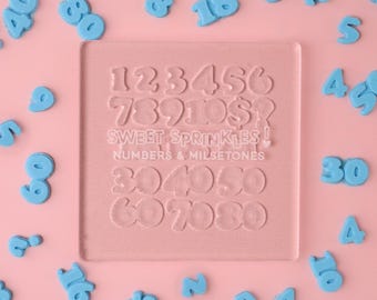 May include: A clear plastic mold with numbers 1 through 80 in a bubble font. The mold is on a pink background with blue plastic numbers scattered around it. The text "SWEET SPRINKLES! NUMBERS & MILESTONES" is printed on the mold.