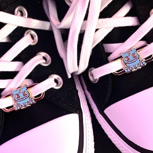 May include: A pair of black canvas shoes with white laces and gold-colored charms featuring a blue cartoon character. The charms are attached to the laces and are visible on both shoes.