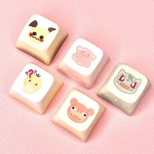 May include: Five square keycaps with cartoon character designs. The keycaps are beige, pink, and gray, with images of a Mimikyu, Ditto, Psyduck, Slowpoke, and Bulbasaur. The keycaps are arranged on a pink surface.
