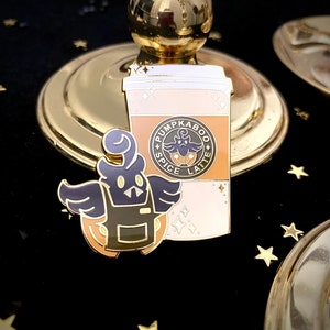 May include: Gold enamel pin featuring a cartoon ghost wearing a brown apron and holding a pumpkin. The ghost is flying above a coffee cup with the text "Pumpkin Spice Latte" on the side.