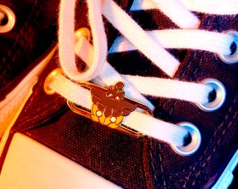 May include: Close-up of a black canvas sneaker with white laces and a gold-colored charm. The charm features a brown and yellow design. The sneaker has a white rubber toe cap and metal eyelets.
